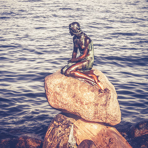 Foto einer Bronzefigur einer Meerjungfrau, die auf einem Stein am Wasser sitzt.