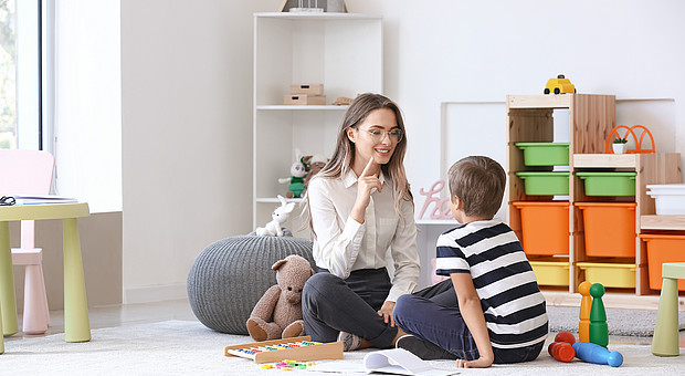 Kind und Erwachsene sitzen auf dem Boden eines hellen Zimmers mit Spielzeug und Regalen im Hintergrund.