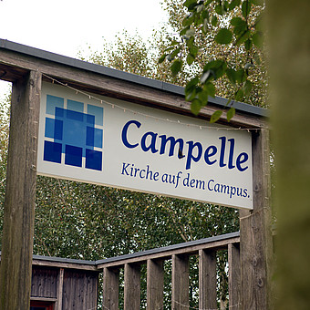 zeigt ein hängendes Schild zwischen Bäumen mit der blauen Aufschrift "Campelle: Kirche auf dem Campus" 