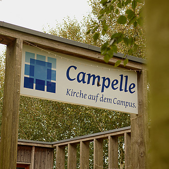 zeigt ein hängendes Schild zwischen Bäumen mit der blauen Aufschrift "Campelle: Kirche auf dem Campus" 