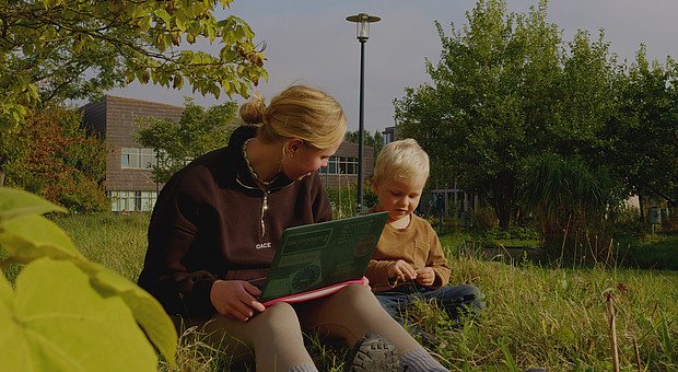 eine Studierende mit Laptop und ein kleines Kind sitzen auf einer Wiesen und entdecken die Natur 