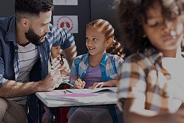 eine Lehrkraft und ein lächelndes Mädchen das im Unterricht am Tisch arbeitet