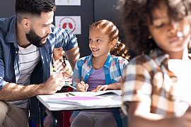 eine Lehrkraft und ein lächelndes Mädchen das im Unterricht am Tisch arbeitet