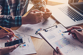 Mehrere Personen sitzen an einem Tisch mit Laptop, Smartphone, Brille und Dokumenten mit Diagrammen und Grafiken darauf.