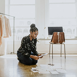 Eine junge BiPoC Frau betrachtet ihre erstellten Mode Designs auf Papier in einem Hellen Raum 