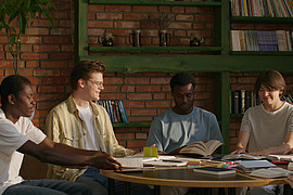 eine gemischte Gruppe Studierender sitzt an einem Tisch und diskutiert Fachliteratur 