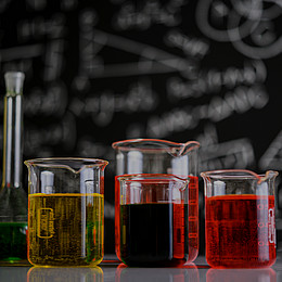 Sieben Laborgläser mit unterschiedlichen Farben stehen auf einem Tisch vor einer Tafel mit Formeln. 