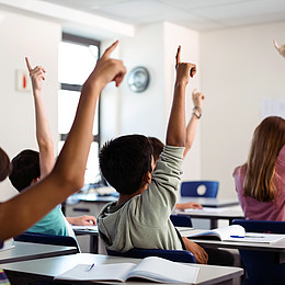 Ein Klassenzimmer in einer Grundschule. Zu sehen sind fünf Schulkinder mit erhobener Hand.  