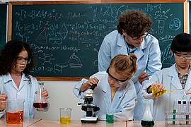 zeigt eine Lehrkraft mit drei Kindern, beim Assistieren eines Experiments in Laborkitteln und Schutzbrillen 