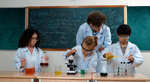 zeigt eine Lehrkraft mit drei Kindern, beim Assistieren eines Experiments in Laborkitteln und Schutzbrillen 