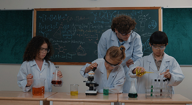 zeigt eine Lehrkraft mit drei Kindern, beim Assistieren eines Experiments in Laborkitteln und Schutzbrillen 