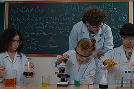 zeigt eine Lehrkraft mit drei Kindern, beim Assistieren eines Experiments in Laborkitteln und Schutzbrillen 