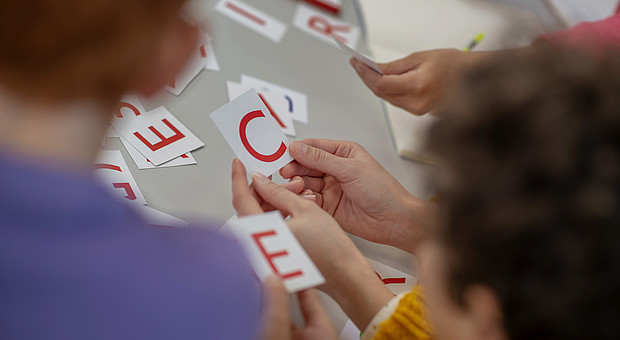 Eine Gruppe von Kindern hält verschiedene Buchstaben hoch. Im Hintergrund ist eine Lehrkraft zu sehen. 