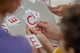 Eine Gruppe von Kindern hält verschiedene Buchstaben hoch. Im Hintergrund ist eine Lehrkraft zu sehen. 