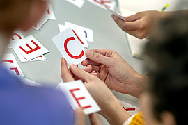 Eine Gruppe von Kindern hält verschiedene Buchstaben hoch. Im Hintergrund ist eine Lehrkraft zu sehen. 