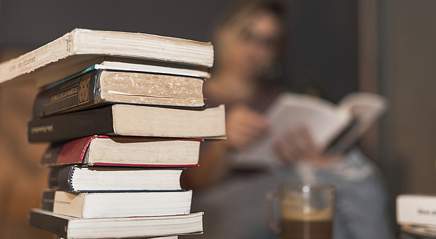 Ein Stapel von Büchern steht mit einer Tasse Kaffee auf einem Wohnzimmertisch. Im Hintergrund ist eine junge Person am Lesen. 