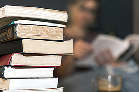 Ein Stapel von Büchern steht mit einer Tasse Kaffee auf einem Wohnzimmertisch. Im Hintergrund ist eine junge Person am Lesen. 