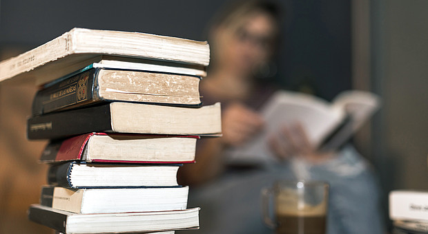 Ein Stapel von Büchern steht mit einer Tasse Kaffee auf einem Wohnzimmertisch. Im Hintergrund ist eine junge Person am Lesen. 