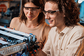 Zwei Studierende mit Schutzbrille an einem Tisch. Sie führen gemeinsam ein Experiment zur Robotik durch.