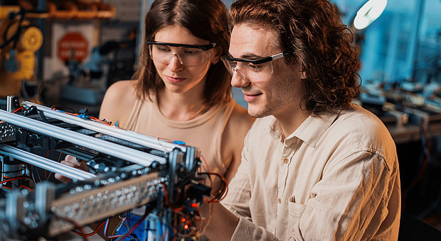 Zwei Studierende mit Schutzbrille an einem Tisch. Sie führen gemeinsam ein Experiment zur Robotik durch.