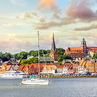 Ein weißes Segelschiff im Hafen der Stadt Flensburg. Im Hintergrund historische Gebäude am Wasser.