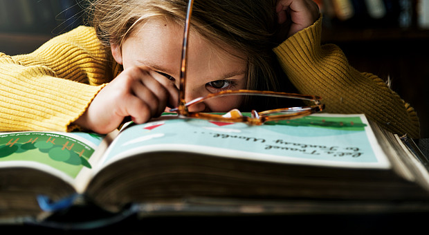 Nahaufnahme eines kleinen Mädchens vor einem dicken Buch. Eine Braune Brille liegt auf dem Buch. 