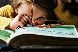 Nahaufnahme eines kleinen Mädchens vor einem dicken Buch. Eine Braune Brille liegt auf dem Buch. 