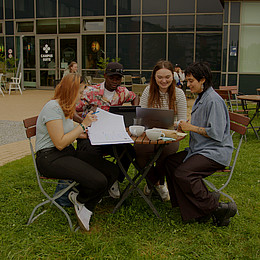 Vier Studierende sitzen draußen auf dem Campus der Europa-Universität Flensburg, trinken Kaffee und lernen. Es sind Kaffeetassen und Laptos auf dem Tisch. 