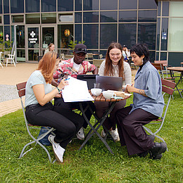 Vier Studierende sitzen draußen auf dem Campus der Europa-Universität Flensburg, trinken Kaffee und lernen. Es sind Kaffeetassen und Laptos auf dem Tisch. 