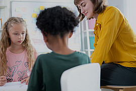 eine Lehrkraft und zwei junge Schüler*innen sitzen an einem kleinen weißen Tisch und machen Aufgaben 