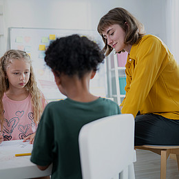 eine Lehrkraft und zwei junge Schüler*innen sitzen an einem kleinen weißen Tisch und machen Aufgaben 