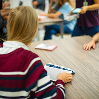 Schulklasse sitzt an Tischen, eine Schülerin mit gestreiftem Pullover hält ein Tablet, im Hintergrund stehen und sitzen weitere Schüler*innen.