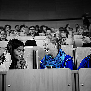 Bildausschnitt aus einem Hörsaal, auf dem sich zwei Frauen unterhalten und lachen.