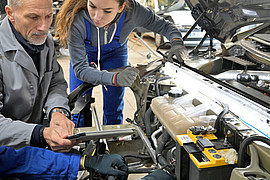 Drei Personen in Arbeitskleidung prüfen gemeinsam den Motorraum eines Fahrzeugs in einer Werkstatt.