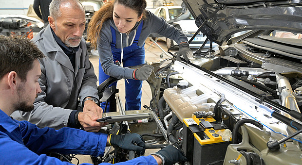 Drei Personen in Arbeitskleidung prüfen gemeinsam den Motorraum eines Fahrzeugs in einer Werkstatt.