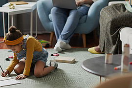 ein kleines Mädchen in Latzhose sitzt mit Stift und Papier auf dem Teppich und malt 