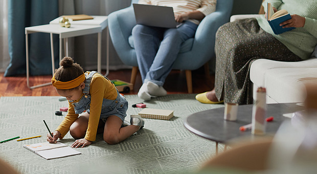 ein kleines Mädchen in Latzhose sitzt mit Stift und Papier auf dem Teppich und malt 