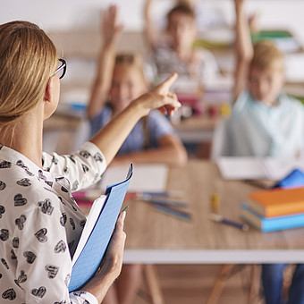 Lehrerin mit Brille und Zopf zeigt mit ausgestrecktem Arm auf einen Schüler, der im Klassenzimmer an einem Tisch sitzt und die Hand hebt.