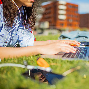 Studentin schreibt auf einem Laptop bäuchlings liegend auf der Wiese