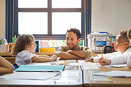 Grundschulkinder sitzen an Tischen in einem Klassenzimmer und schreiben oder zeichnen auf Papier, zwischen ihnen ein lächelnder Lehrer.