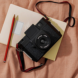 eine Kamera, ein Bleistift und ein Notizblock liegen auf einem roten Laken 