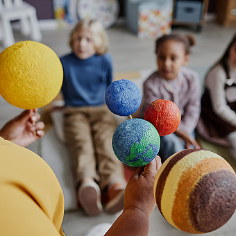 Nahaufnahme eines Planetenmodells in der Hand einer Lehrkraft vor sitzenden Kindern 