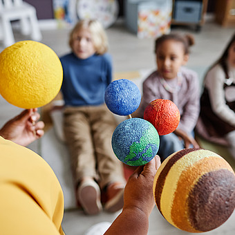 Nahaufnahme eines Planetenmodells in der Hand einer Lehrkraft vor sitzenden Kindern 