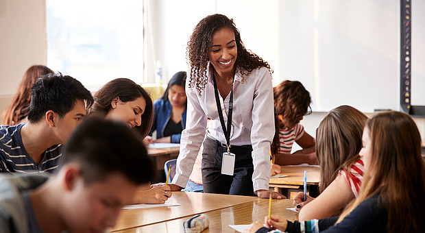 Lehrerin steht an einem Tisch und unterrichtet eine Gruppe von Schüler*innen, die an Tischen sitzen und schreiben.