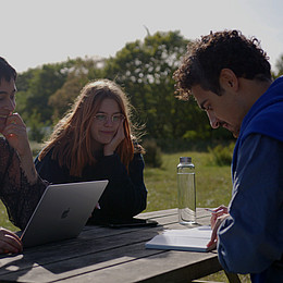 drei Studierende sitzen am Ostseebad von Flensburg und lernen bei gutem Wetter an einem Holztisch 