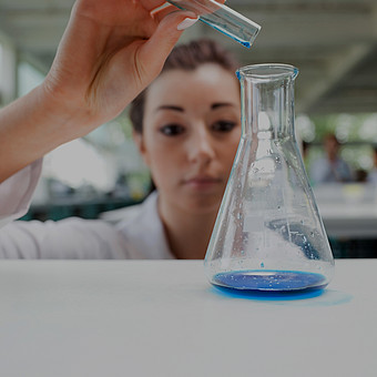 Eine Chemie Studierende im Laborkittel gießt eine blaue Flüßigkeit aus ihrem Reagenzglas in ein Laborglas.