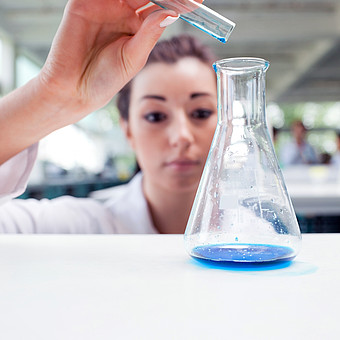 Eine Chemie Studierende im Laborkittel gießt eine blaue Flüßigkeit aus ihrem Reagenzglas in ein Laborglas.