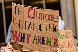 Menschen bei einer Klimademonstration halten Pappschilder hoch; auf einem steht: „The climate is changing, why aren’t you?“