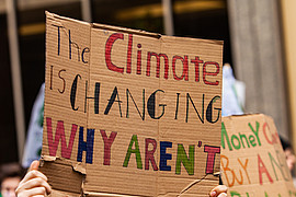 Menschen bei einer Klimademonstration halten Pappschilder hoch; auf einem steht: „The climate is changing, why aren’t you?“