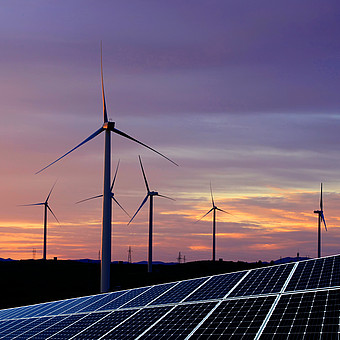 Several wind turbines and an area with solar panels stand in a hilly landscape. In the background, a picturesque sunset with a violet-orange sky.